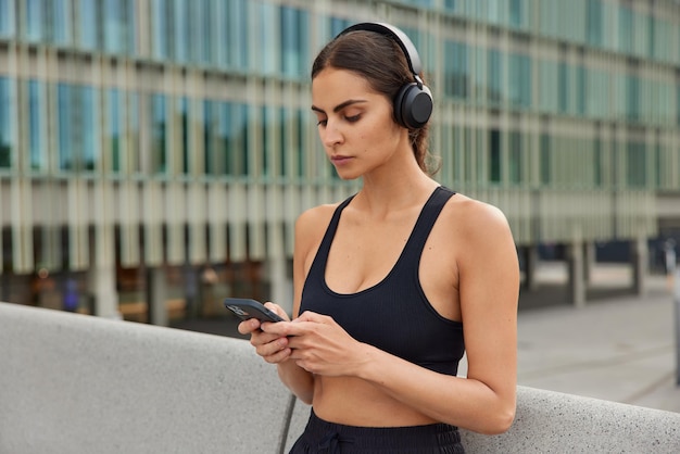 la donna concentrata sul display dello smartphone vestita con abbigliamento sportivo indossa cuffie wireless sulle orecchie posa contro l'edificio della città