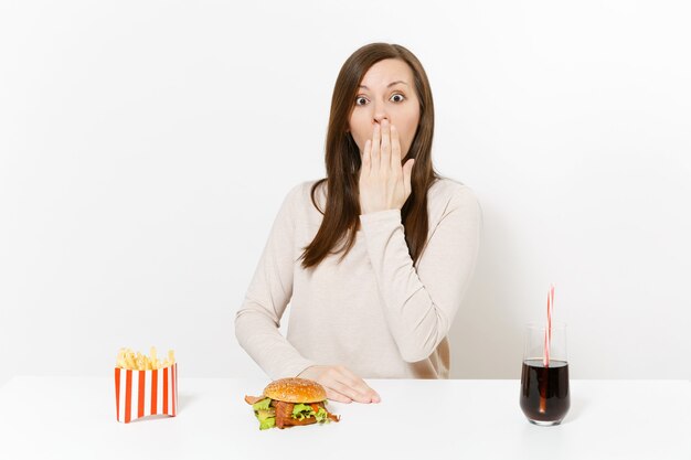 La donna copre la bocca con la mano al tavolo con hamburger, patatine fritte, cola in bottiglia di vetro isolata su sfondo bianco. Una corretta alimentazione o un classico fast food americano. Area pubblicitaria con copia spazio.