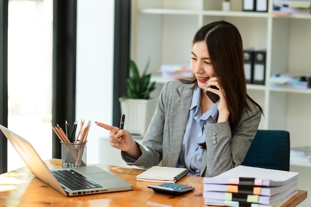 La donna d'affari asiatica felice ha la gioia di parlare sul tablet smartphone e sul laptop nell'ufficio moderno