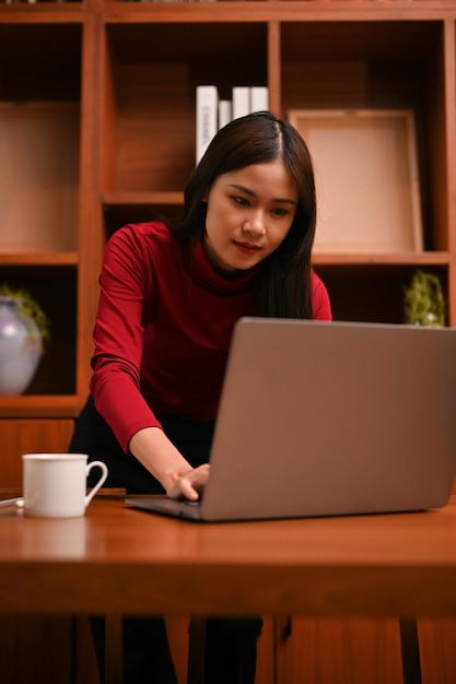 La donna d'affari asiatica in piedi si appoggiò alla scrivania concentrandosi sul suo progetto sul laptop