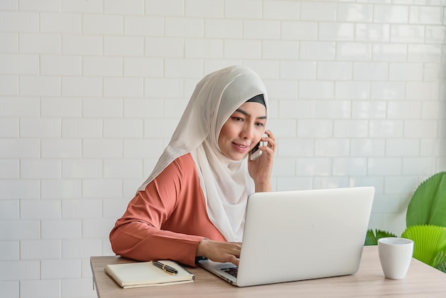 La donna d'affari asiatica musulmana usa il computer portatile e lo smartphone nella caffetteria o nello spazio di coworking