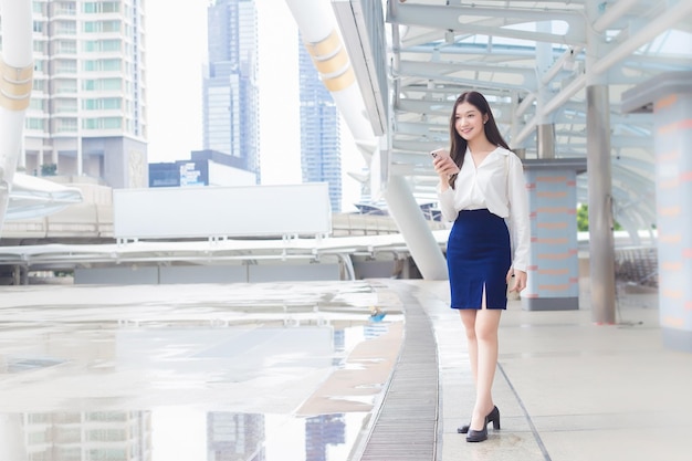 La donna d'affari asiatica sta andando al posto di lavoro dell'ufficio che tiene lo smartphone con la città come sfondo