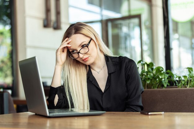 La donna d'affari bionda stanca guarda nello schermo del laptop mentre si siede a un tavolo in un caffè all'aperto Lavoro remoto freelance