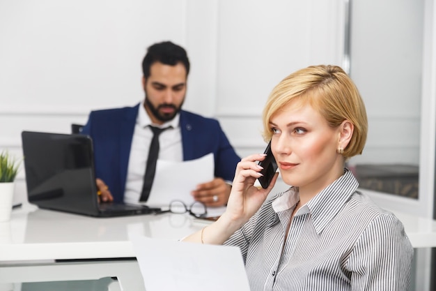 La donna d'affari che lavora ha una conversazione telefonica con l'uomo barbuto che lavora con il computer portatile nell'interno moderno dell'ufficio bianco