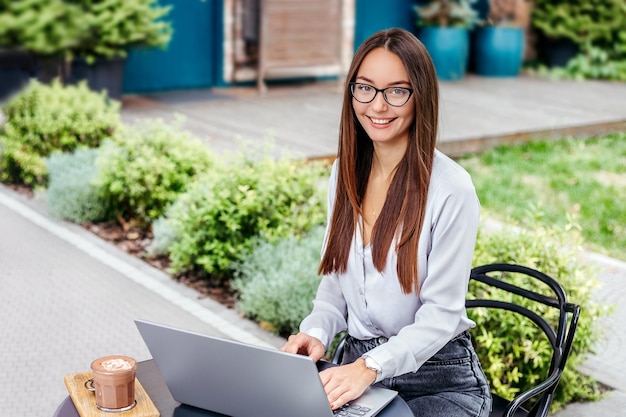 La donna d'affari con gli occhiali usa il laptop sorridendo e guardando la telecamera seduto in un caffè sulla strada