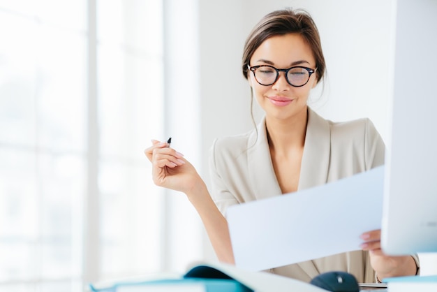 La donna d'affari di successo concentrata guarda attentamente negli studi su carta i termini del contratto tiene la penna scrive nelle documentazioni vestita formalmente posa sul desktop contro interni spaziosi bianchi