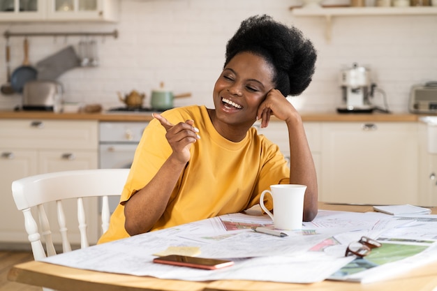 La donna d'affari nera felicissima ride il lavoro da casa architetto allegro si siede al tavolo con i progetti