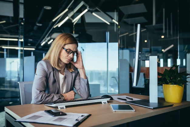 La donna d'affari senior ha un forte mal di testa mentre lavora in un ufficio moderno al computer