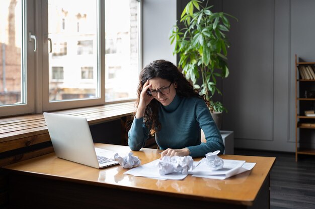 La donna d'affari stanca sopraffatta si siede alla scrivania con il computer portatile e la carta stropicciata frustrata ed esausta