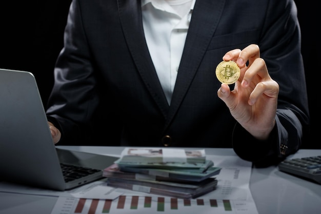 La donna d'affari tiene in mano la moneta reale della criptovaluta Bitcoin per presentare lo spazio della copia del mockup della finanza decentralizzata su sfondo nero con il grafico dei soldi delle banconote del computer portatile sul tavolo