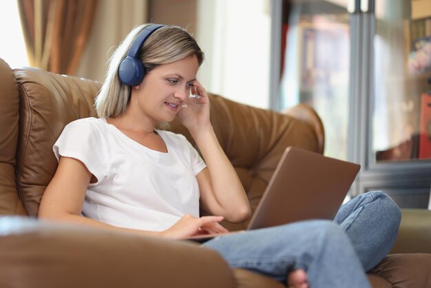 La donna dai capelli biondi lavora al computer portatile e ascolta la musica in cuffia seduta su un comodo divano