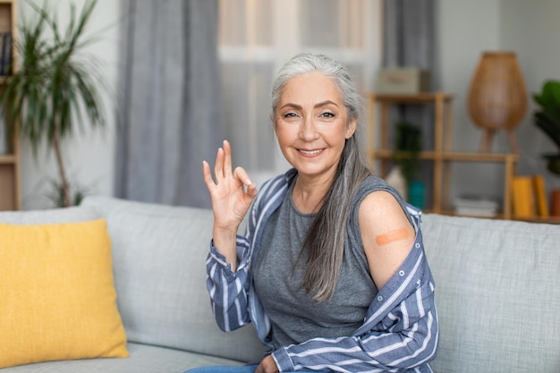La donna dai capelli grigi europea senior sorridente mostra il segno giusto con la mano e la spalla con il cerotto dopo la vaccinazione
