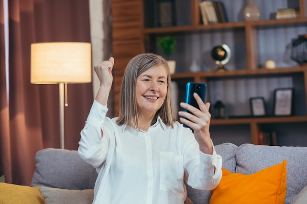 La donna dai capelli grigi senior felice ha ricevuto buone notizie online seduta sul divano di casa e si rallegra celebrando la vittoria guardando lo schermo del telefono