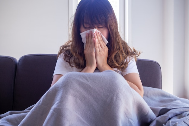 La donna dai capelli lunghi seduta sul divano soffre di influenza, tosse e starnuti. Seduto in una coperta a causa della febbre alta e coprirsi il naso con carta velina perché starnutisce tutto il tempo.