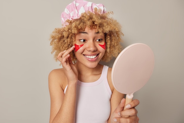 La donna dai capelli ricci allegra si guarda allo specchio mentre applica le macchie rosse di bellezza sotto gli occhi essendo di buon umore indossa il cappello da bagno e una maglietta casual isolata su sfondo grigio Procedure di cosmetologia