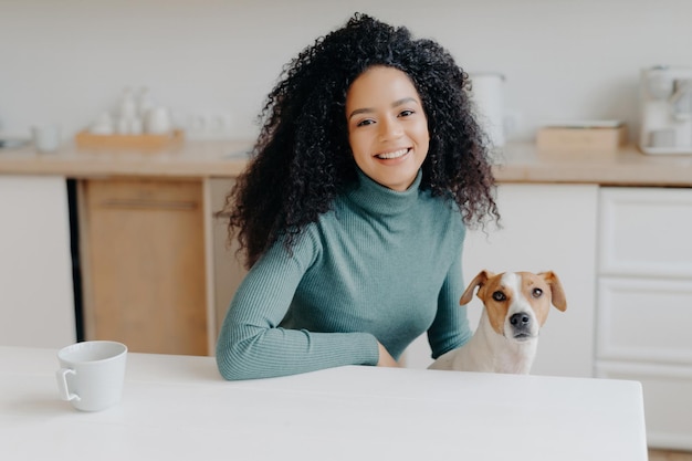 La donna dai capelli ricci in dolcevita si siede al tavolo della cucina e si diverte a giocare a tè con Jack Russell terrier Momento gioioso con l'amato animale domestico