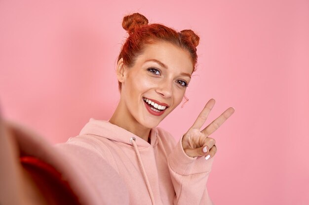 La donna dai capelli rossi con il sorriso in felpa con cappuccio rosa fa il segno della pace si fa selfie tramite telefono sul muro rosa