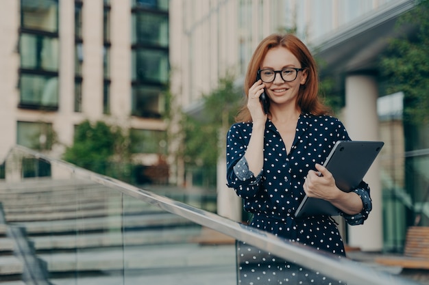 La donna dai capelli rossi in abiti alla moda ha una conversazione telefonica che cammina per strada vicino a un edificio moderno