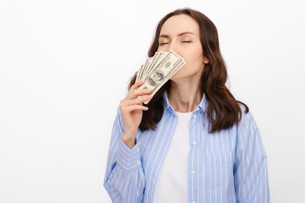 La donna dai capelli scuri in camicia blu annusa i dollari che tiene in mano sul concetto di risparmio su sfondo bianco