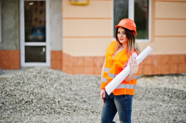 La donna del costruttore dell'ingegnere in panciotto uniforme e casco protettivo arancio tiene il rotolo della carta da disegno di affari contro la nuova costruzione