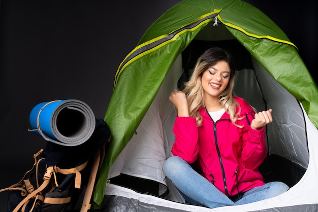 La donna dell'adolescente dentro una tenda verde di campeggio isolata sulla parete nera con i pollici aumenta il gesto e sorridere