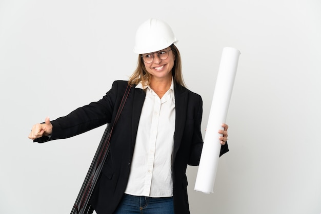 La donna dell'architetto di mezza età con il casco e che tiene le cianografie sopra la parete isolata che dà un pollice aumenta il gesto