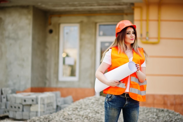 La donna dell'ingegnere in gilet uniforme e casco protettivo arancione tiene il rotolo di carta da disegno aziendale contro il nuovo edificio Tema del blocco vivente della proprietà