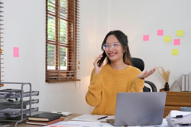 La donna di affari asiatica attraente e sorridente sta parlando su un telefono cellulare con un cliente con il computer portatile nell'ufficio