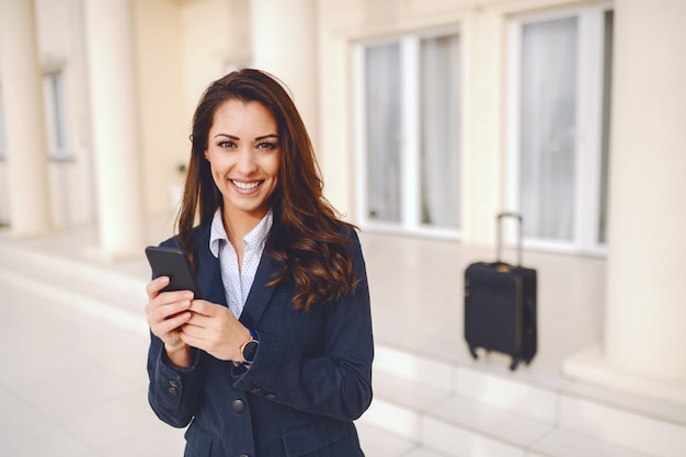 La donna di affari caucasica sorridente dei giovani ha vestito casuale astuto usando astuto per la scrittura del messaggio mentre stava all'aperto. Nel bagaglio di fondo. Concetto di viaggio d'affari.