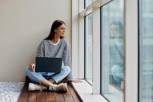 La donna di affari che si siede con un computer portatile alla finestra fulllength e la donna felice sorridente lavora online nello spazio della copia dell'ufficio