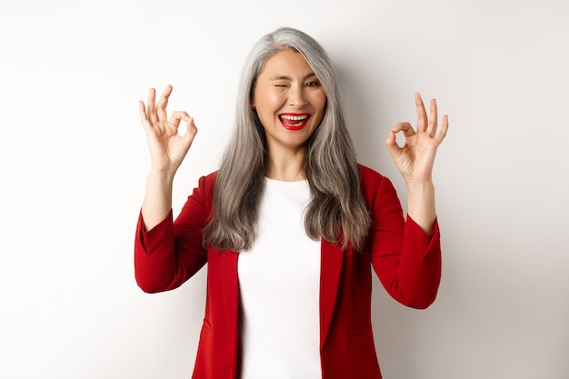 La donna di affari professionale asiatica che mostra i segni giusti e ammiccante, sorridendo compiaciuta, assicura o consiglia qualcosa, levandosi in piedi contro il muro bianco.