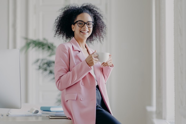 La donna di affari rilassata tiene la tazza della bevanda calda, ha la pausa caffè, sta vicino al suo posto di lavoro in spazioso armadio bianco indossa occhiali lunghi giacca rosa funziona in ufficio. Tempo di riposo dopo il lavoro