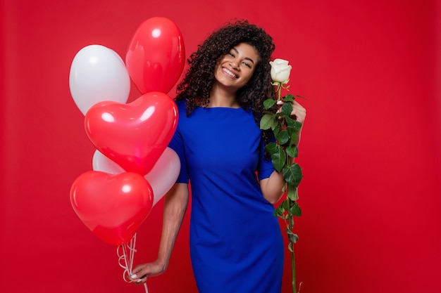 La donna di colore graziosa con la rosa bianca e il cuore ha modellato i palloni variopinti isolati sulla parete rossa