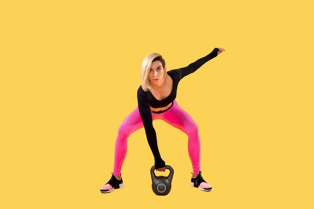 La donna di forma fisica in abiti sportivi alla moda rosa e neri risolve con kettlebell sulla parete gialla. Forza e motivazione.