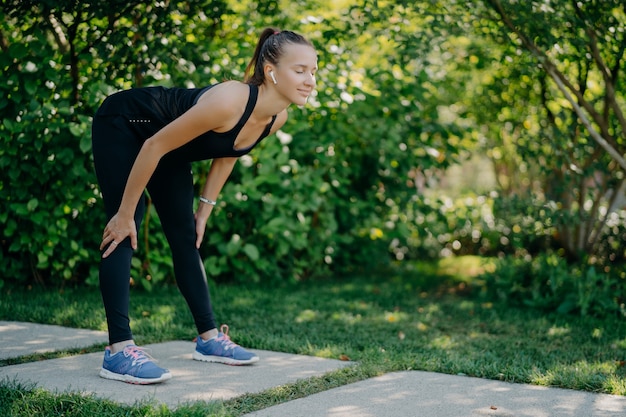 La donna di forma fisica in abiti sportivi neri tiene le mani sulle ginocchia si sporge in avanti esercizi all'aperto contro la natura verde green