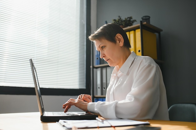 La donna di mezza età matura alla moda sorridente si siede alla scrivania con il ritratto del computer portatile anziana donna d'affari senior