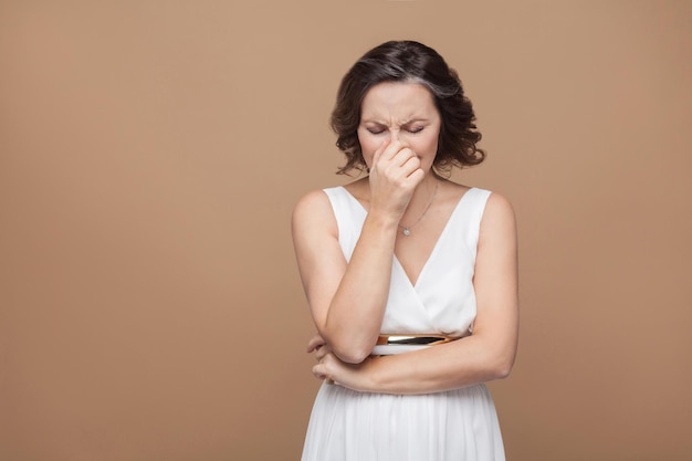 La donna di mezza età triste triste infelice piange nella depressione. Donna che esprime emotiva in abito bianco, labbra rosse e acconciatura riccia scura. Studio girato, indoor, isolato su sfondo beige o marrone chiaro