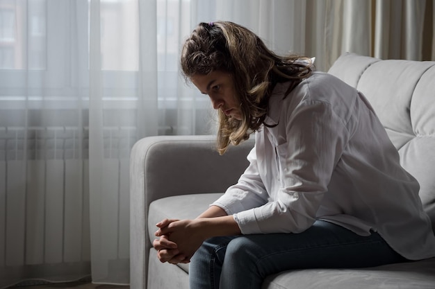 La donna disabile depressa si sente male a causa della paralisi cerebrale infantile seduta sul divano vicino alla finestra nel soggiorno di casa che sperimenta abilismo disabile