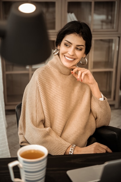 La donna è gentile. Bella signora dai capelli scuri con orecchini dorati e ampio sorriso seduta in maglione lavorato a maglia beige