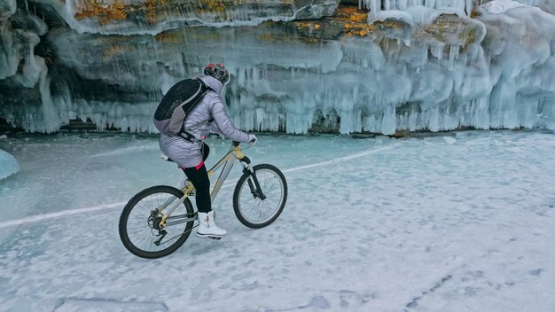 La donna è in bicicletta vicino alla grotta di ghiaccio La roccia con grotte di ghiaccio e ghiaccioli La ragazza è vestita con un piumino argentato zaino in bicicletta e casco Pneumatici invernali con punte Il viaggiatore è in bicicletta