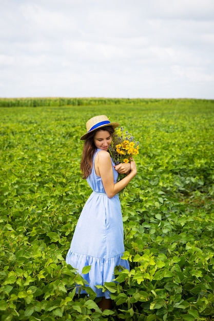 La donna è in piedi nel campo con un bouquet di fiori gialli e un cappello di paglia