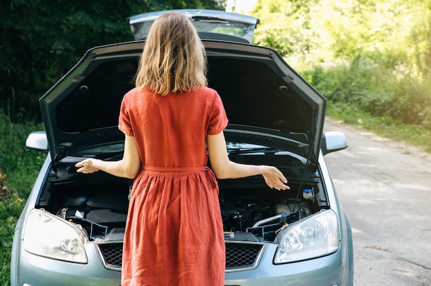 La donna è in piedi vicino all'auto rotta nella natura. Situazione di incidente durante il viaggio. La ragazza vestita non sa cosa fare. Aprire il cofano del veicolo.