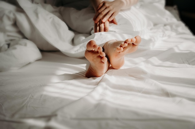 La donna è sdraiata a piedi nudi a letto dormendo sotto lenzuola bianche comode e comode Piedi femminili sul letto Buongiorno donna concetto di sessualità cura di sé e relax concetto di riposo