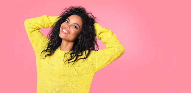 La donna etnica africana con capelli ondulati neri come il jet in felpa lavorata a maglia gialla è in posa mentre guarda la telecamera e sorride con gioia