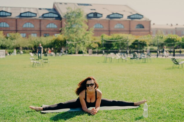 La donna europea attiva in forma fa pilates sul tappetino fitness indossa occhiali da sole e abbigliamento sportivo mostra grandi progressi pose sul prato verde ha treni di espressione felice all'esterno. Concetto di fitness e stretching