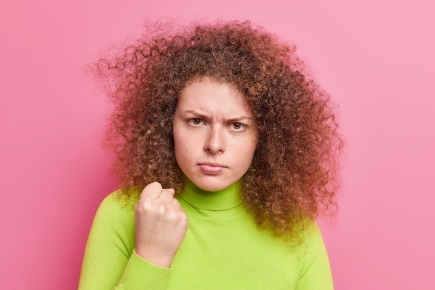 La donna europea dai capelli ricci arrabbiata e infastidita guarda con espressione scontrosa stringe i gesti del pugno incazzato promesse di vendetta vestita casualmente isolata sul muro rosa. concetto di rabbia