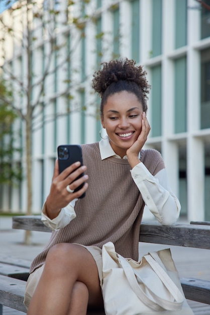 la donna fa clic su se stessa la foto scatta una foto tramite il telefono cellulare sorride piacevolmente si siede all'aperto vicino alla borsa in tessuto utilizza l'applicazione wireless per la comunicazione in roaming