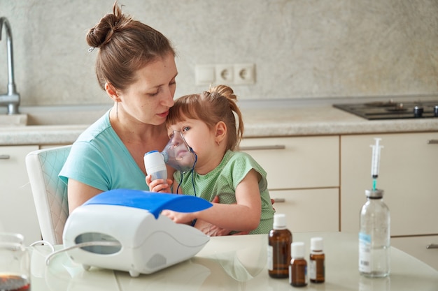 La donna fa l'inalazione a un bambino a casa. porta la maschera del nebulizzatore in faccia. inala il vapore del farmaco. la ragazza respira attraverso la maschera. medicina sul tavolo.