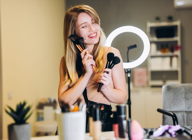 La donna felice con i capelli biondi tiene in mano molti pennelli per il trucco. Bionda che ride con i pennelli per il trucco vicino al viso.