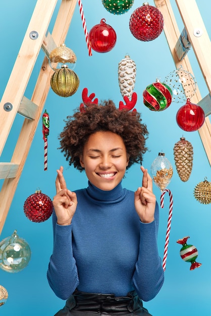 La donna felice con i capelli ricci tiene gli occhi chiusi incrocia le dita crede nella buona fortuna fa pose di desiderio su sfondo blu decora la casa prima di Capodanno. Concetto di persone e vacanze invernali